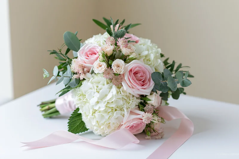 Bridal bouquet with pink roses and white hydrangeas arranged with greenery and ribbon on a white surface