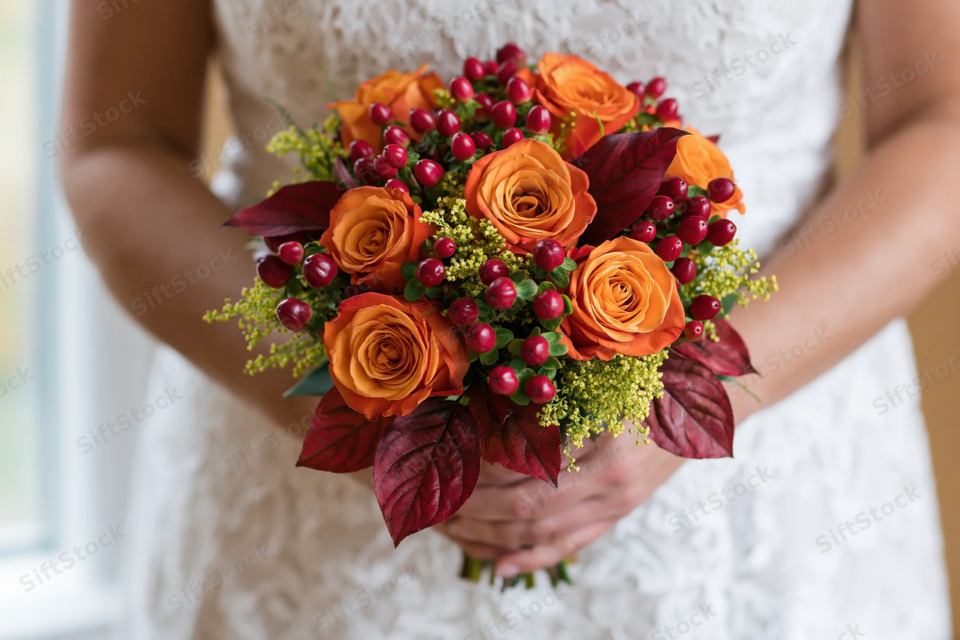 Bride holding autumn wedding bouquet with orange roses and red berries in white lace dress