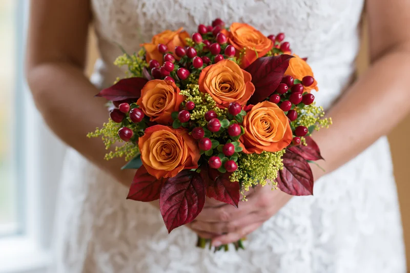 Bride holding autumn wedding bouquet with orange roses and red berries in white lace dress