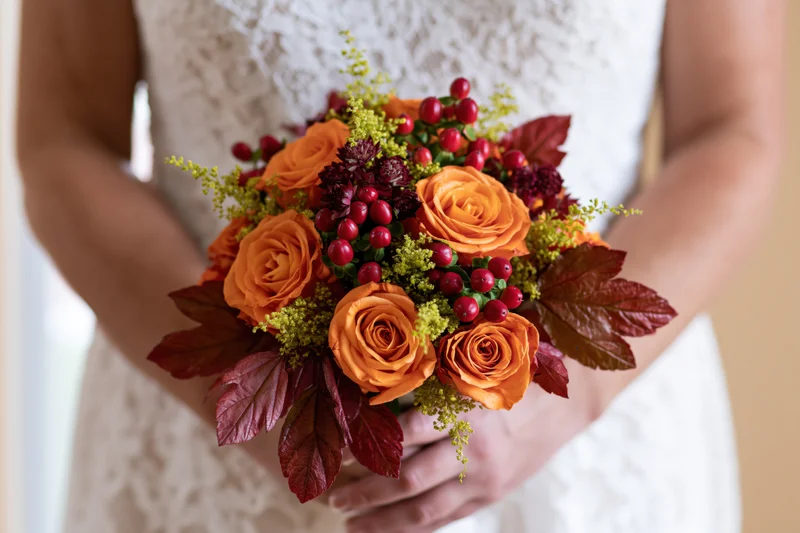 Bride holding autumn wedding bouquet with orange roses and red berries in white lace dress