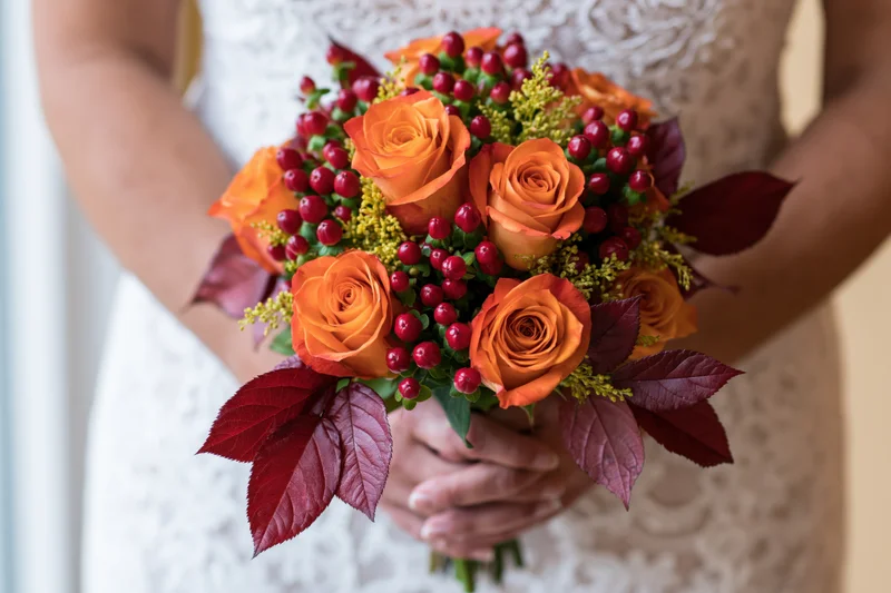 Bride holding autumn wedding bouquet with orange roses and red berries in white lace dress