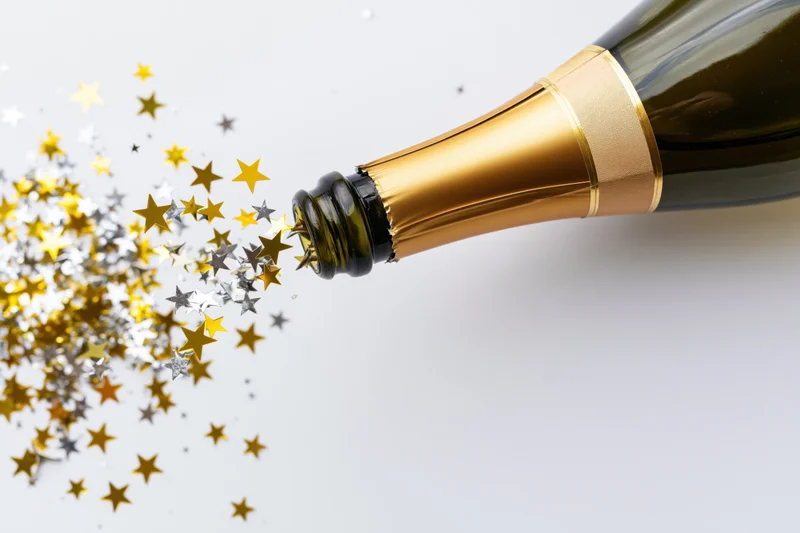 Champagne bottle tilted with gold and silver star confetti spraying out against white background