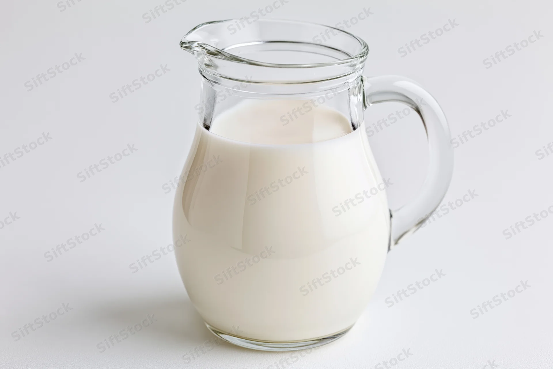 Clear glass pitcher filled with fresh whole milk on white surface