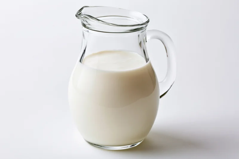 Clear glass pitcher filled with fresh whole milk on white surface
