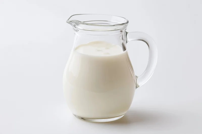 Clear glass pitcher filled with fresh whole milk on white background