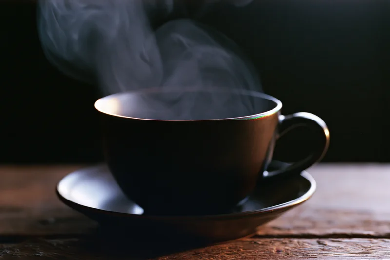 Steaming dark ceramic coffee cup on matching saucer resting on wooden table