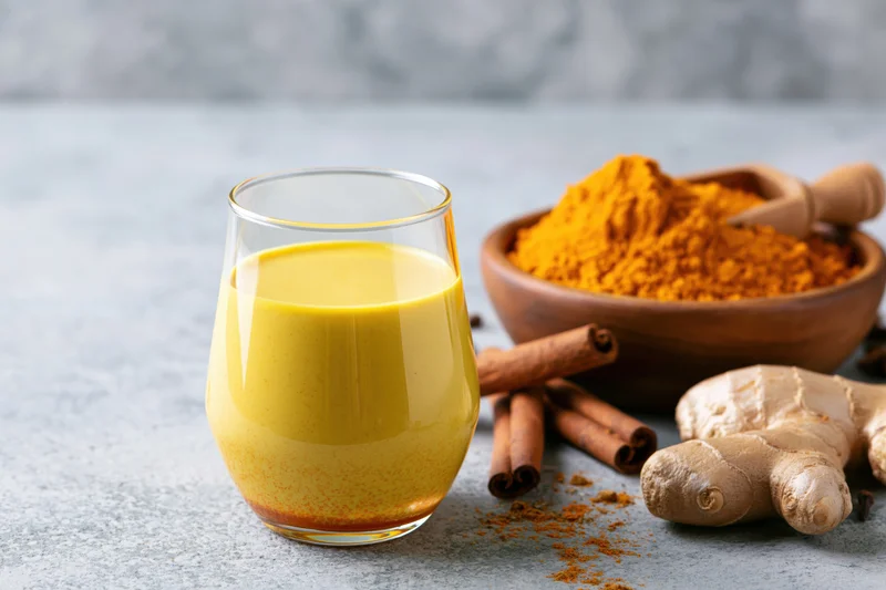 Glass of golden turmeric milk with cinnamon sticks, ginger root, and turmeric powder in a wooden bowl on a gray countertop