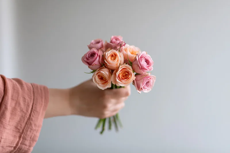 Hand holding a small bouquet of pink and peach roses against a plain background