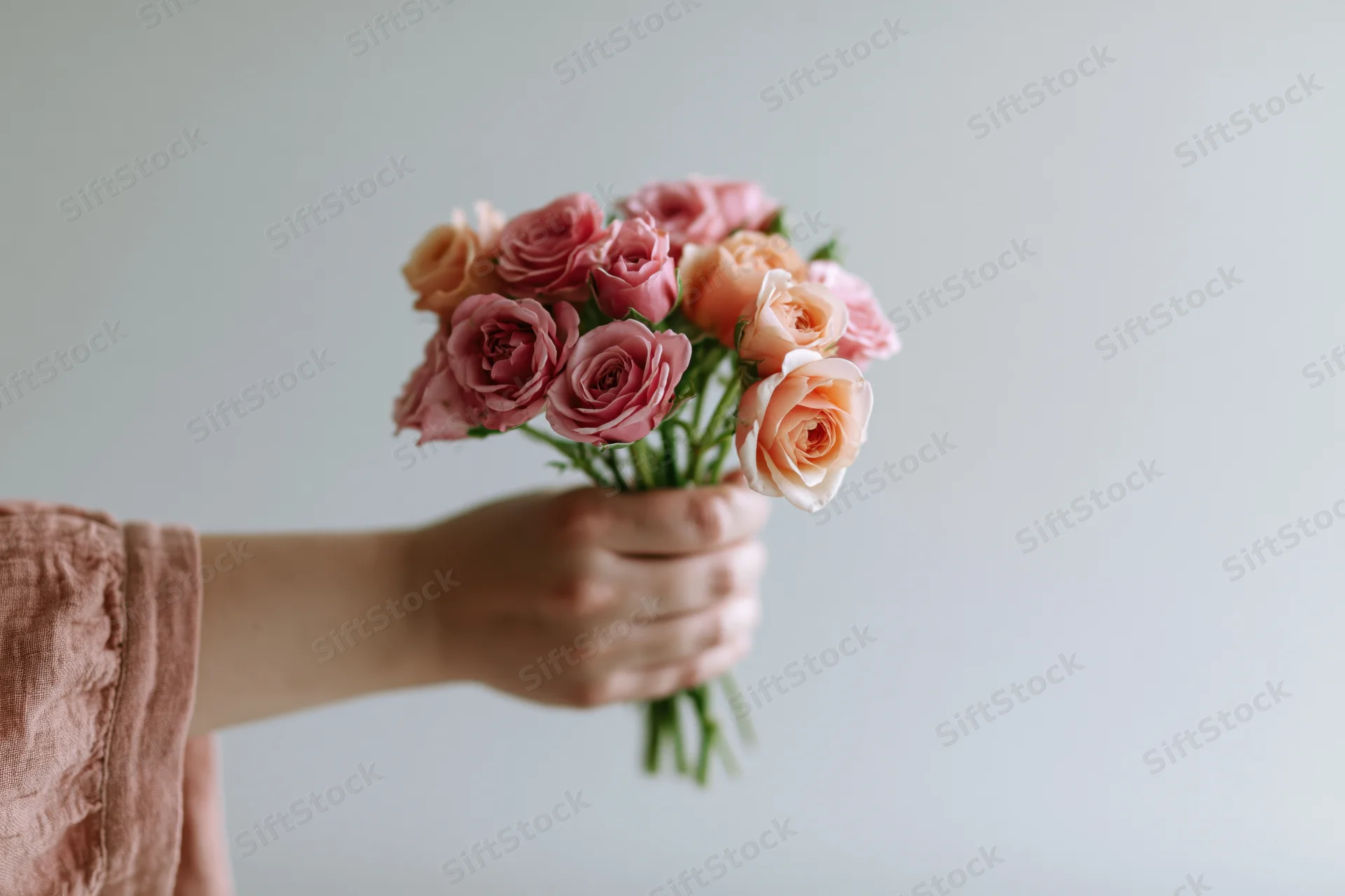 Hand holding a small bouquet of pink and peach roses against a light background