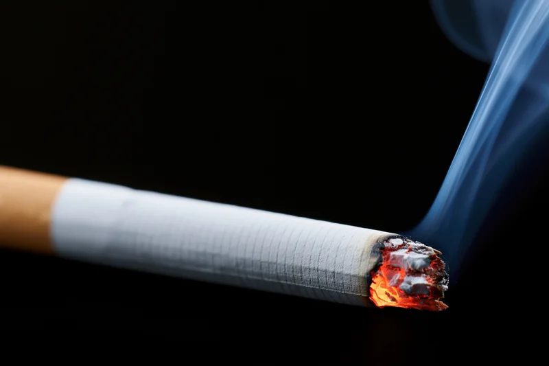 Lit cigarette burning with glowing ember and rising smoke against black background