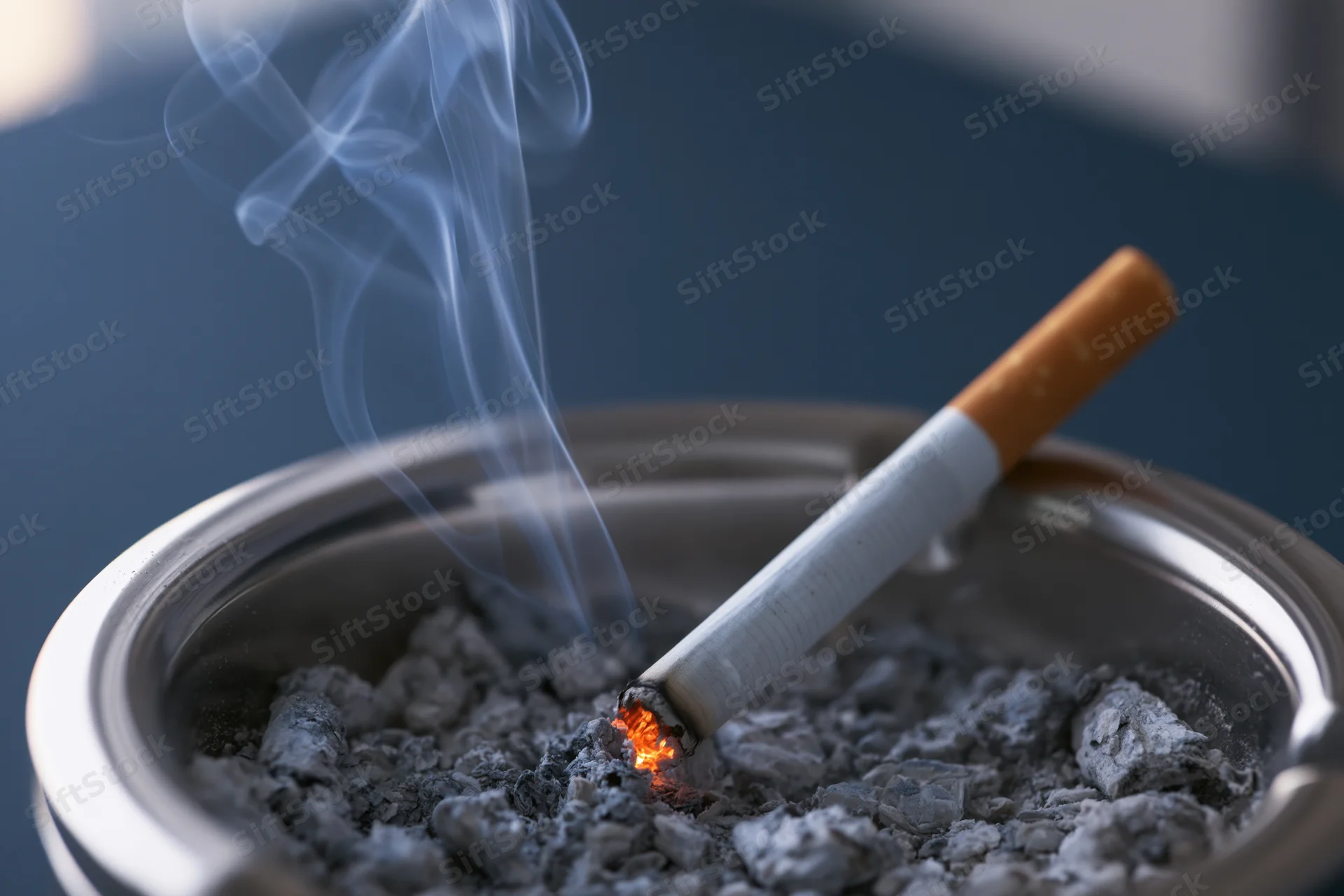 Lit cigarette burning in a metal ashtray filled with ash, smoke rising, close-up view