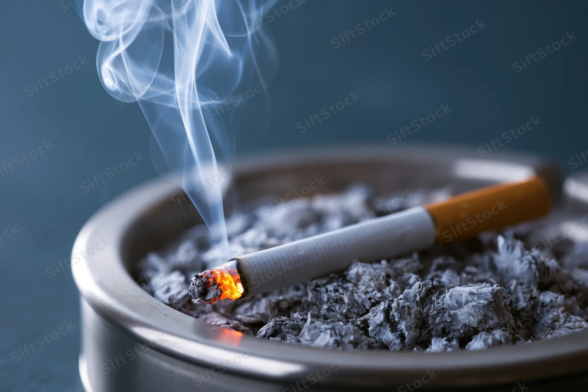 Lit cigarette burning in a metal ashtray filled with ash, emitting smoke