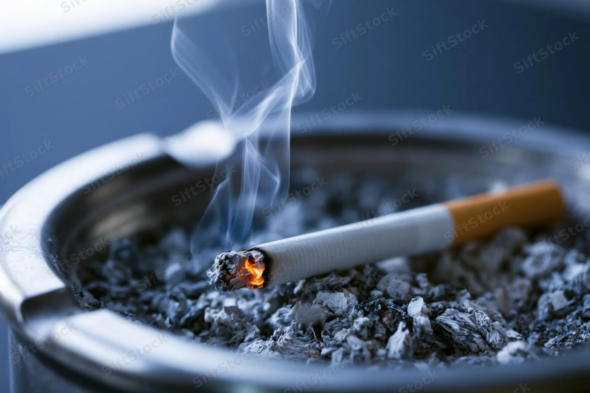 Lit cigarette burning in a metal ashtray filled with ash, close-up view
