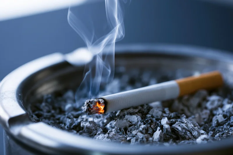Lit cigarette burning in a metal ashtray filled with ash, close-up view