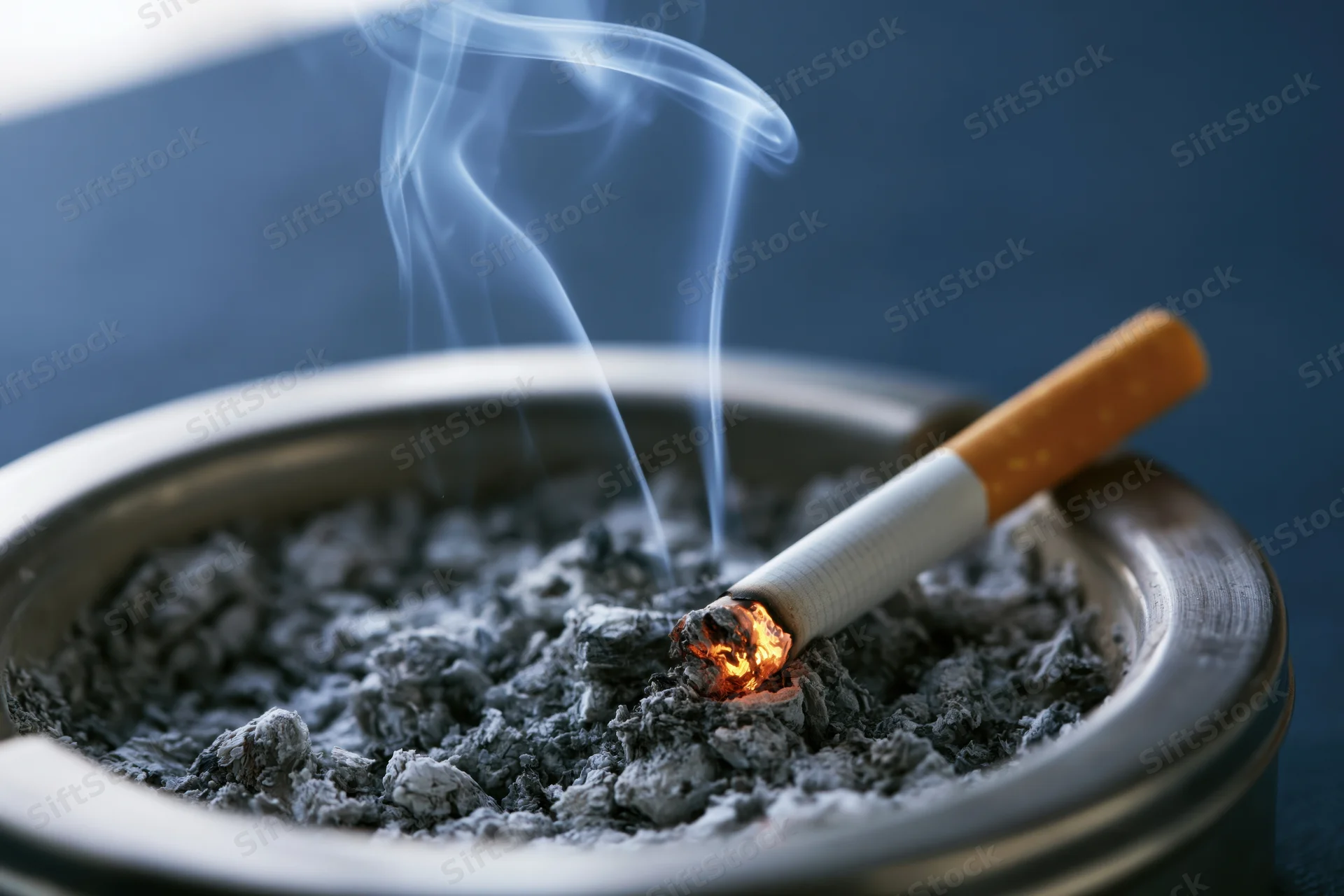 Lit cigarette burning in a metal ashtray filled with ash, emitting smoke
