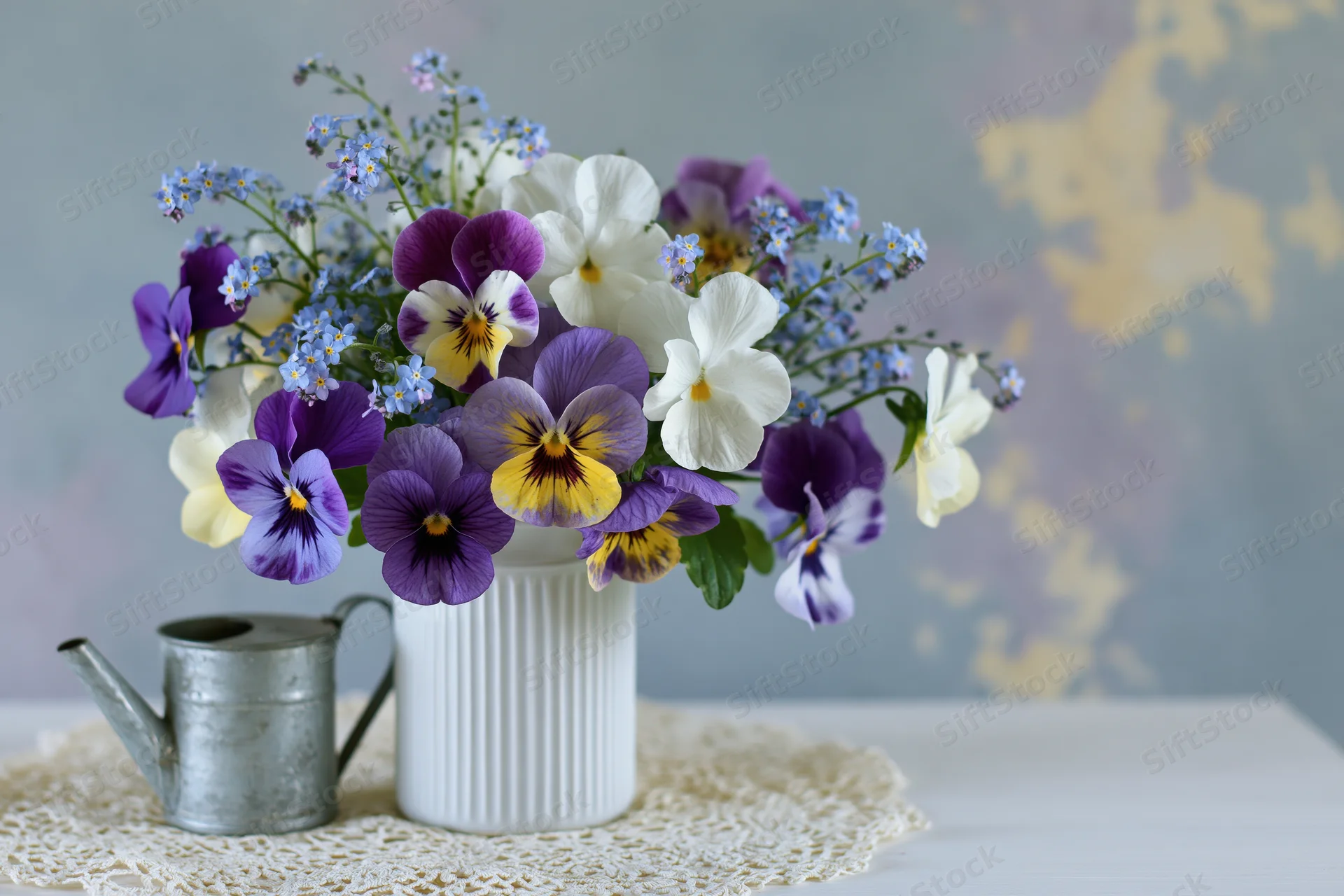 A loose bouquet of purple, white, and yellow pansies with forget-me-nots in a white ribbed vase on a lace doily beside a metal watering can on a table