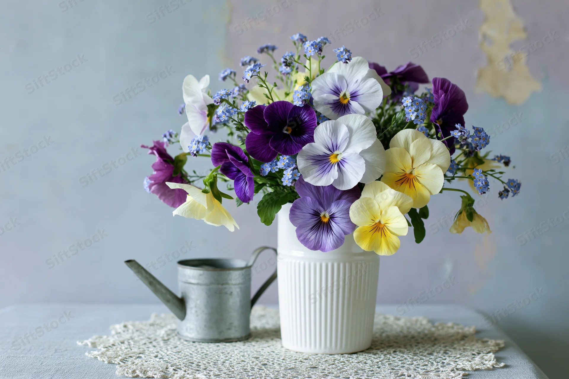 A loose bouquet of purple white and yellow viola pansies in a white ribbed vase on a lace doily with a metal watering can beside it on a table