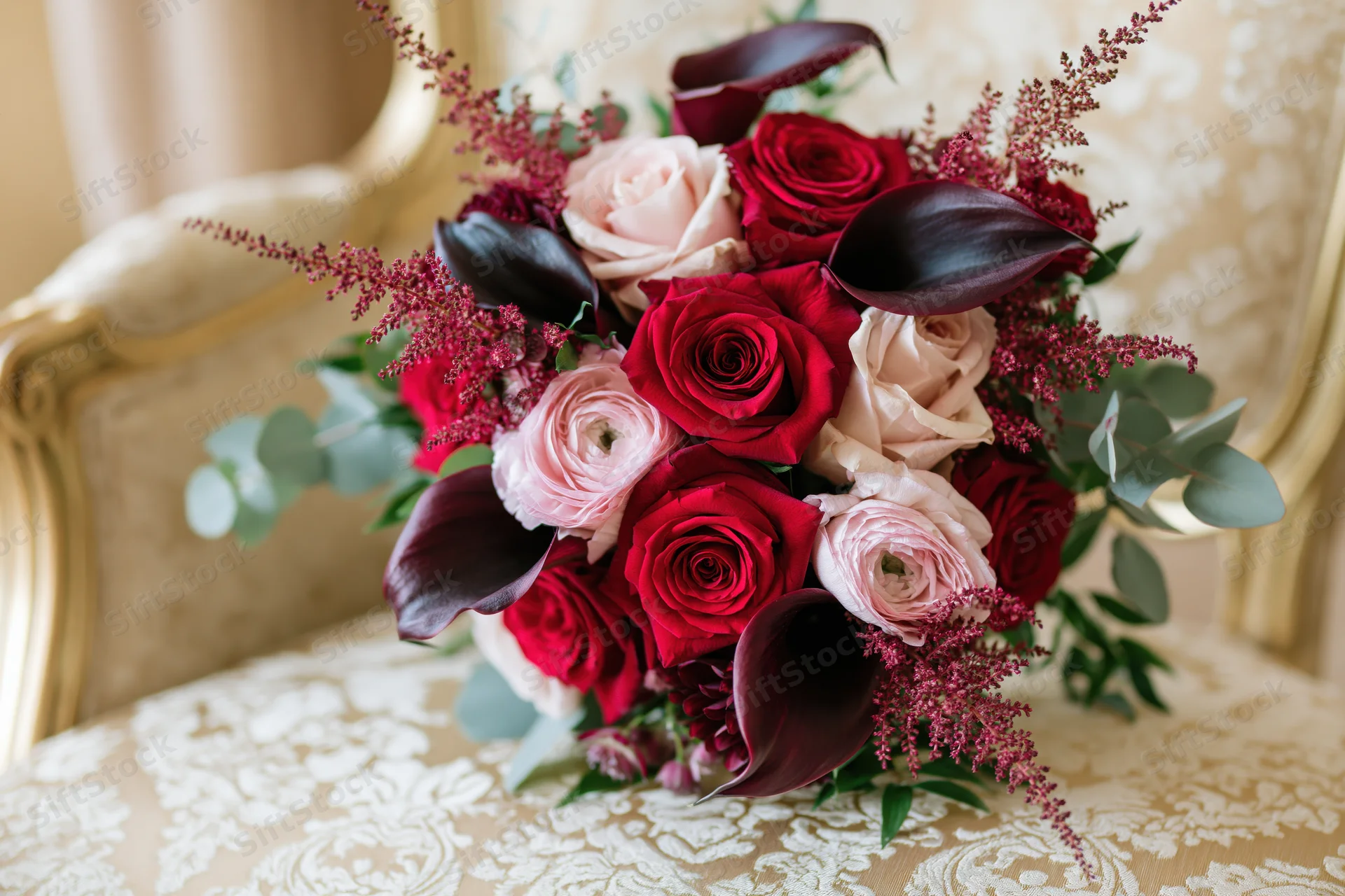Bridal bouquet with red and pink roses and dark calla lilies resting on ornate cream and gold upholstered chair