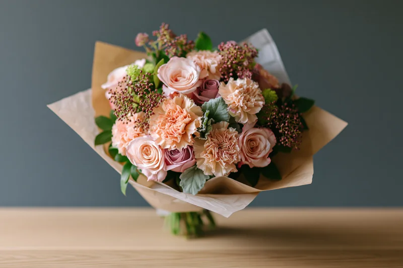 Mixed bouquet of blush roses and peach carnations wrapped in kraft paper on wooden table