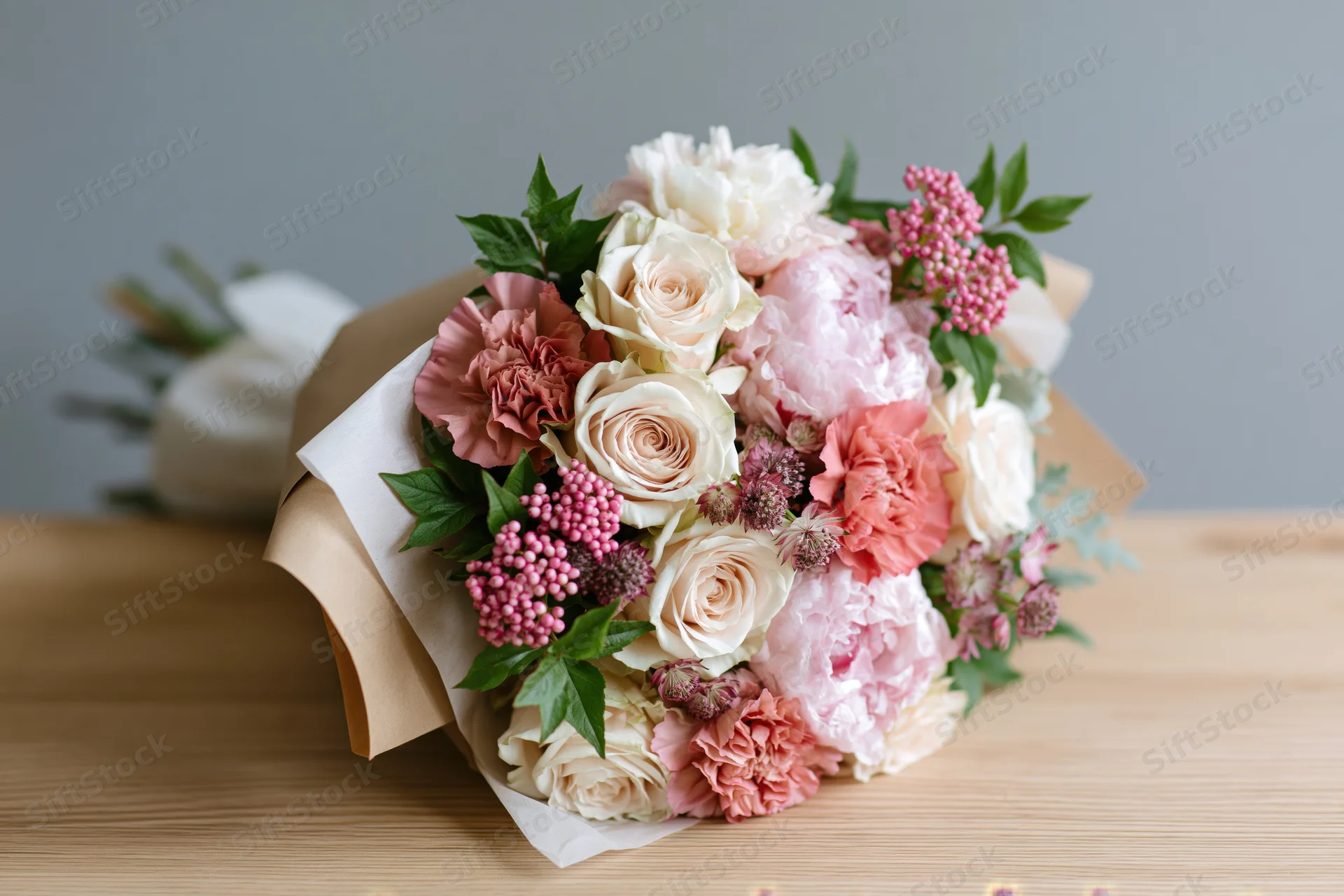 Mixed bouquet of cream roses and blush pink peonies with carnations and greenery wrapped in kraft paper on wooden table