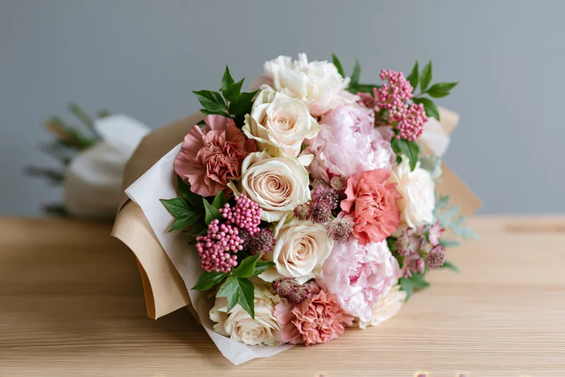 Mixed bouquet of cream roses and blush pink peonies with carnations and greenery wrapped in kraft paper on wooden table