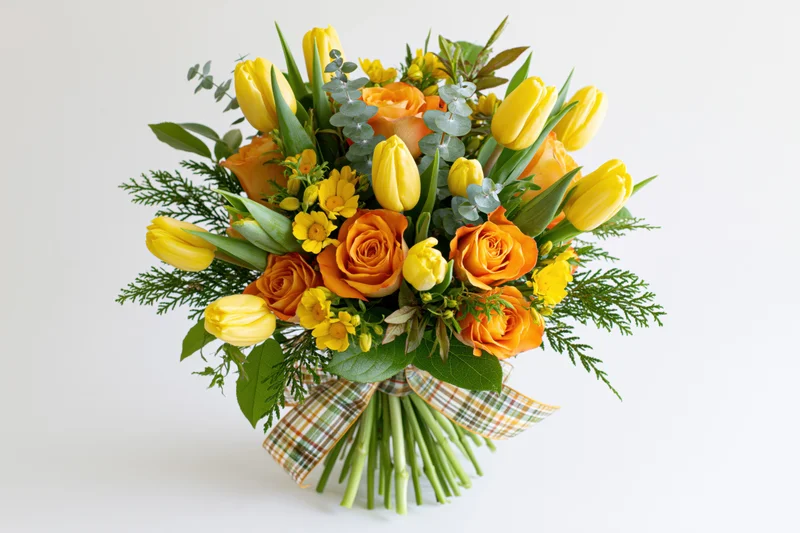 Mixed bouquet of yellow tulips and orange roses with greenery and plaid ribbon on white background