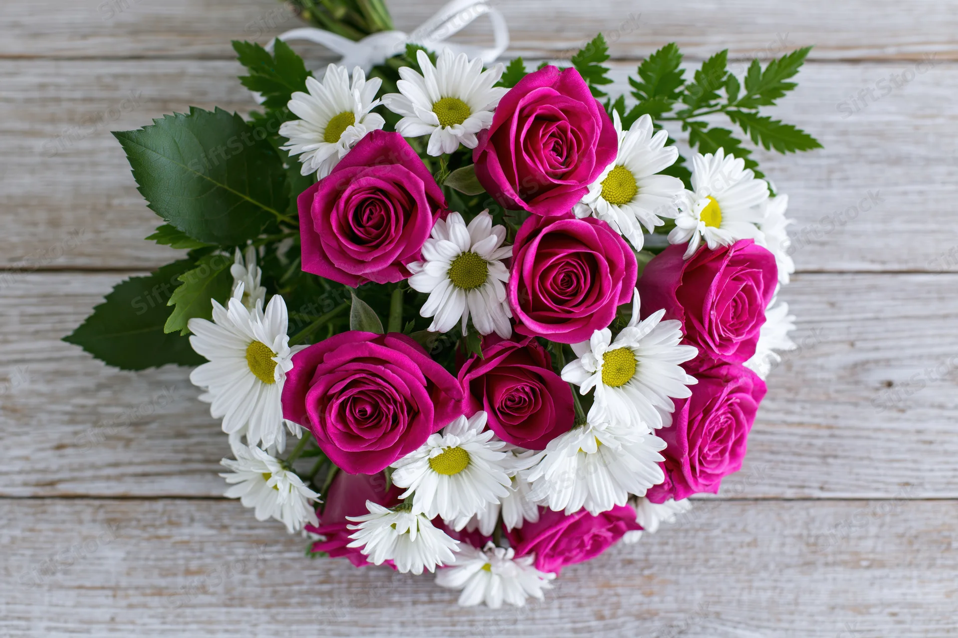 Mixed bouquet of deep pink roses and white daisies with green foliage on a rustic wooden surface