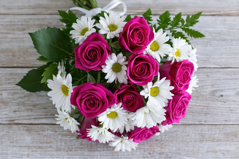 Mixed bouquet of deep pink roses and white daisies with green foliage on a rustic wooden surface