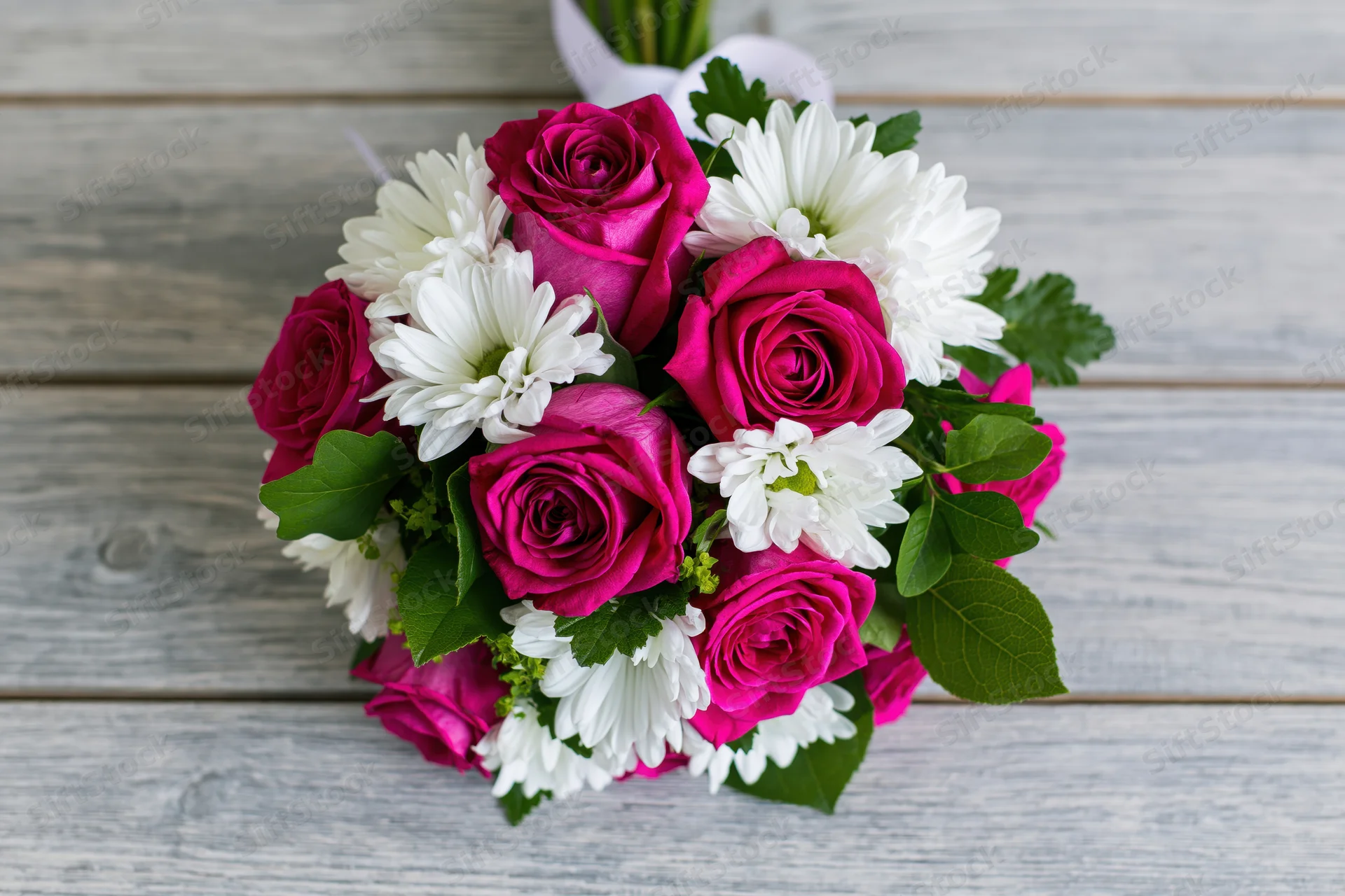 A bouquet of deep pink roses and white daisies with green leaves arranged on a rustic wooden table