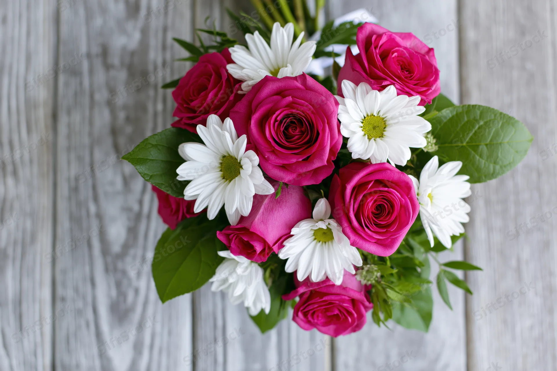 A bouquet of deep pink roses and white daisies with green leaves arranged on a wooden surface