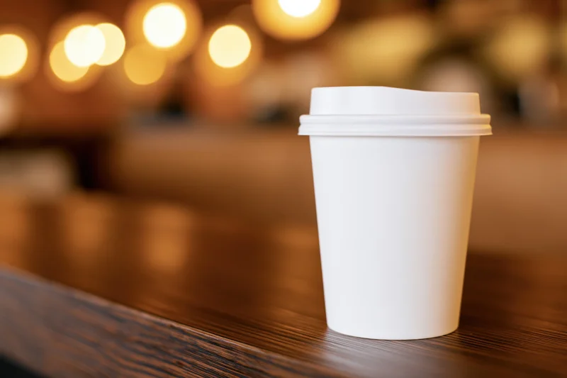 White paper coffee cup on wooden table in cafe interior with warm bokeh lights in background