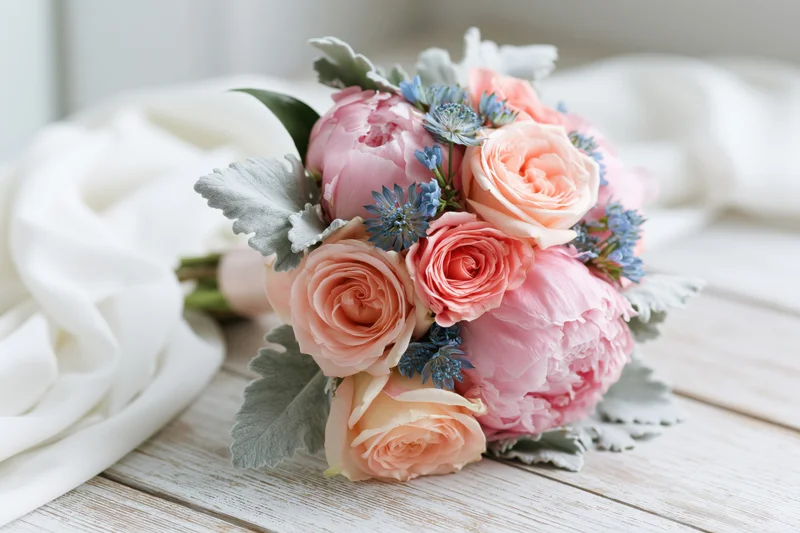 Round bridal bouquet of pink peonies and peach garden roses with blue flowers and silver foliage on a wooden table