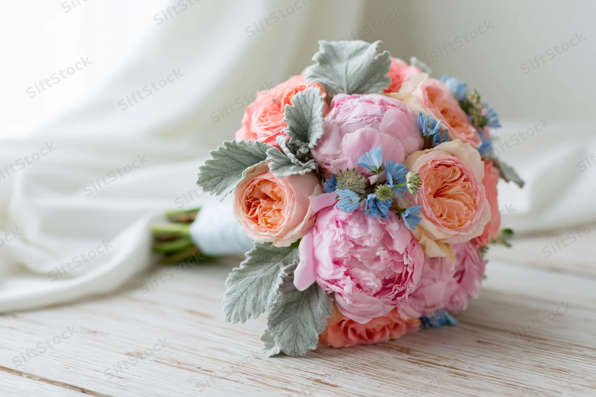 Round bridal bouquet of pink peonies and peach roses with blue flowers and silver foliage on wooden table