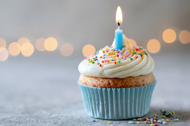 Single birthday cupcake with lit candle and sprinkles in blue paper liner on textured surface with bokeh lights behind