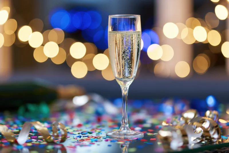 Champagne flute filled with sparkling wine on a table scattered with confetti and streamers, with blurred festive lights in the background