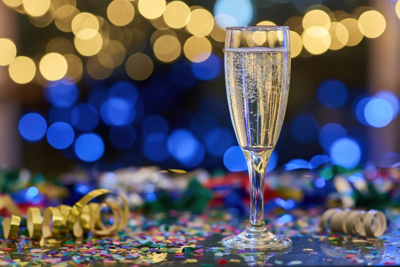Champagne flute filled with sparkling wine on a table covered in confetti and streamers with blurred festive lights in the background