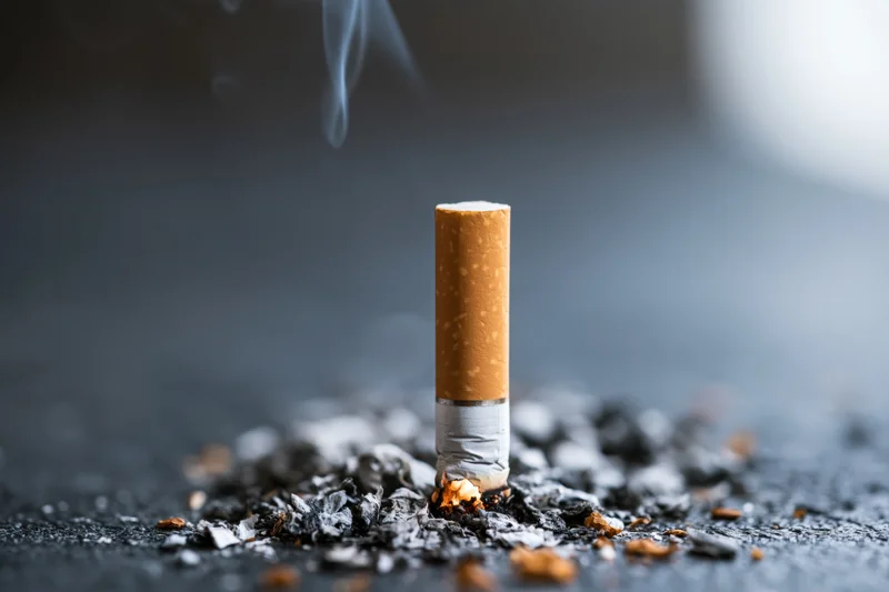 Cigarette butt standing upright on dark surface with smoke rising and ash scattered around