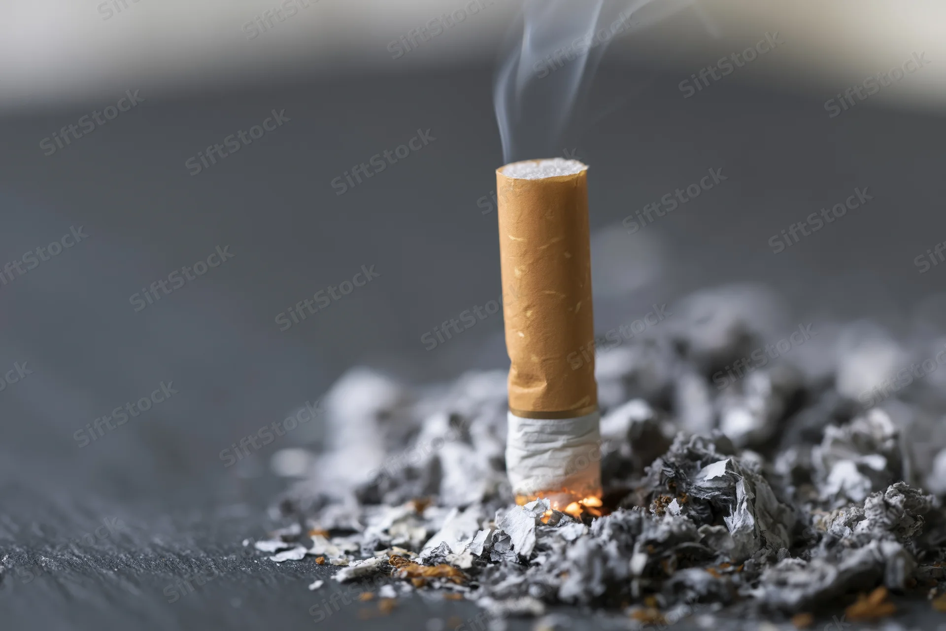 Cigarette butt with smoke rising from ember on dark surface surrounded by ash