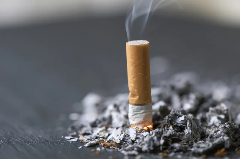 Cigarette butt with smoke rising from ember on dark surface surrounded by ash