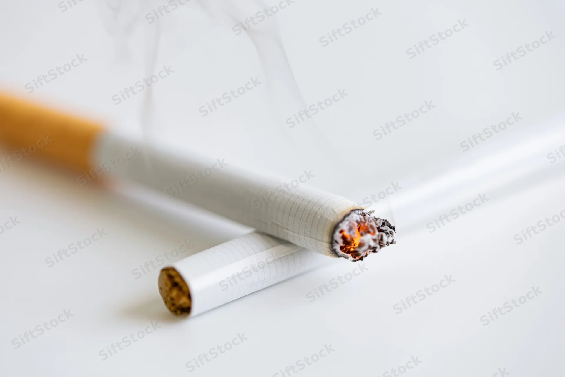 Lit cigarette burning with smoke rising, resting diagonally on white surface