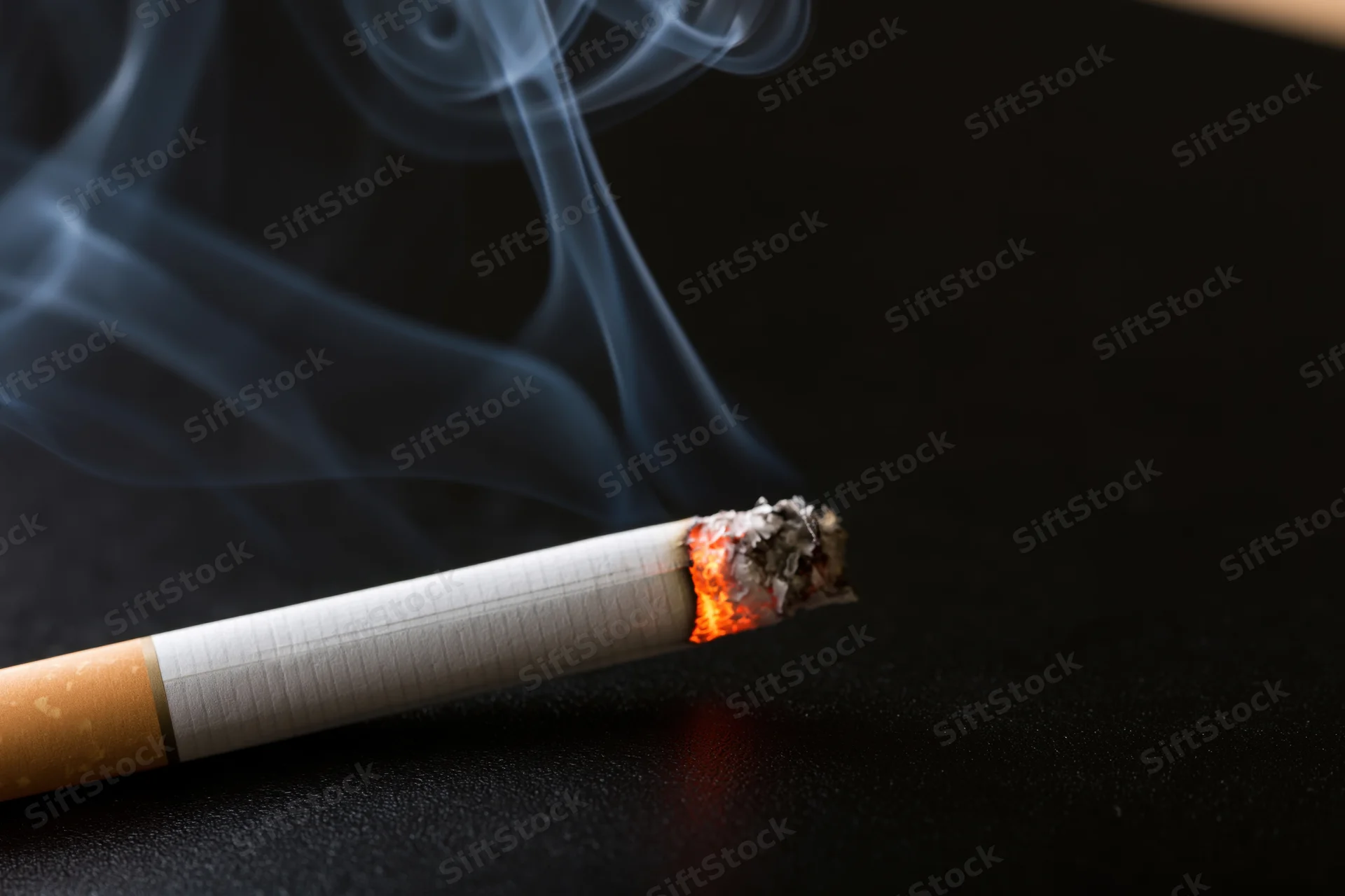 Lit cigarette burning with smoke rising on dark surface