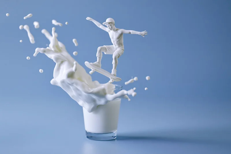 White milk sculpture of a child skateboarding emerging from a glass of milk against a blue background