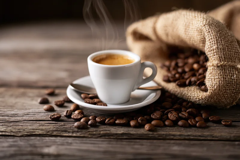 Steaming espresso cup with crema on saucer beside burlap sack of coffee beans on wooden table