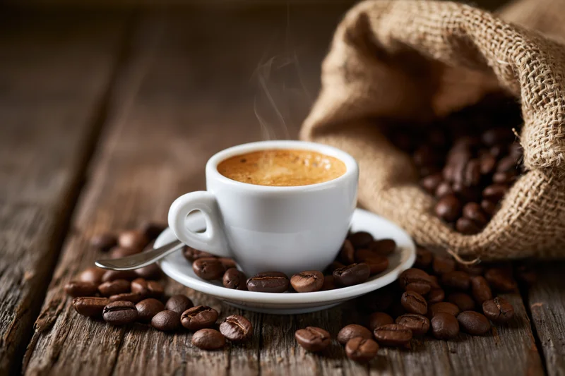 Steaming espresso cup with crema on saucer surrounded by coffee beans on wooden table near burlap sack
