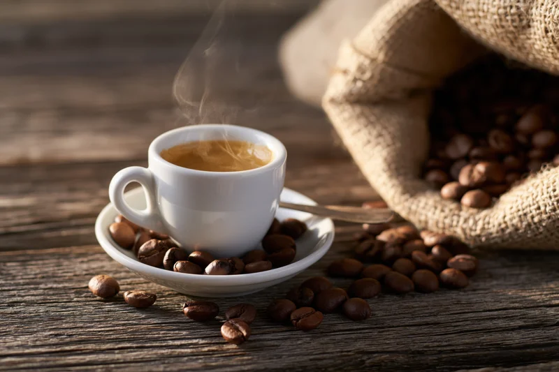 Steaming espresso cup with crema on saucer surrounded by coffee beans on wooden table near burlap sack