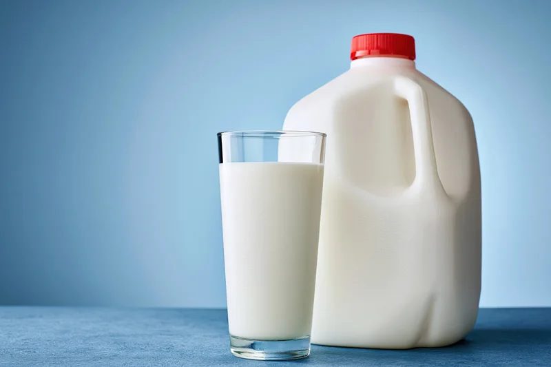 Tall glass of whole milk beside a plastic jug on a blue surface