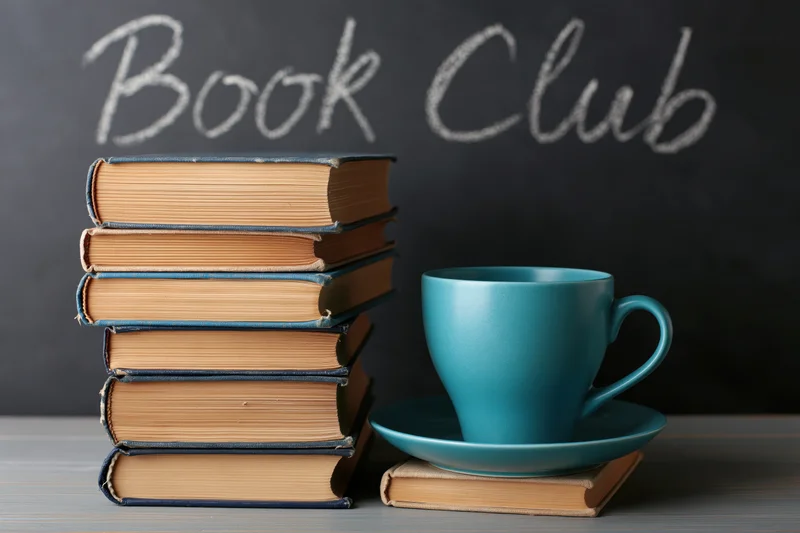 Stack of worn hardcover books beside a teal ceramic mug on a table with book club written in chalk on a blackboard behind