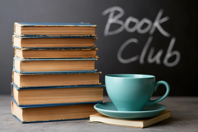 Stack of worn hardcover books beside a teal ceramic cup on a gray surface with book club text on a chalkboard background