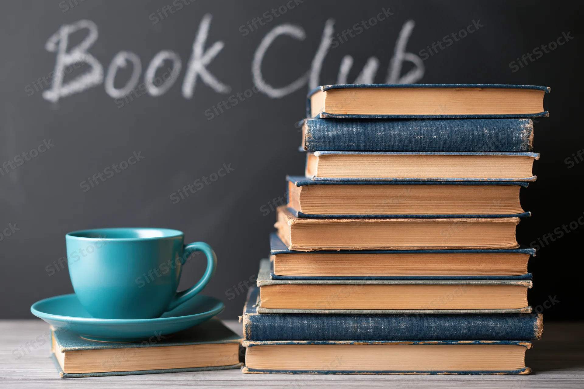Stack of worn hardcover books beside a teal ceramic mug on a wooden table with book club written in chalk on a blackboard behind