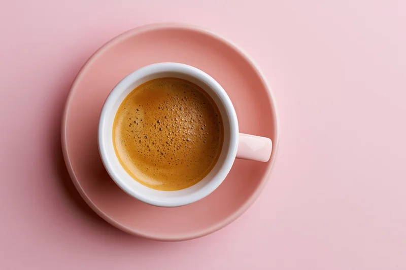 White ceramic coffee cup with frothy espresso on pink saucer, overhead view